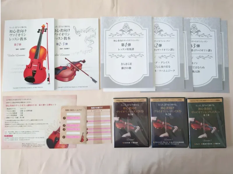 初心者向けヴァイオリンDVDの同梱内容の写真(箱から出して並べた状態)・解説用DVD 3セット ・テキスト教本 2個(2・3弾共通) ・レッスン用楽譜 3個 ・ピアノ伴奏譜 2個 ・指板シール・台紙 各1枚